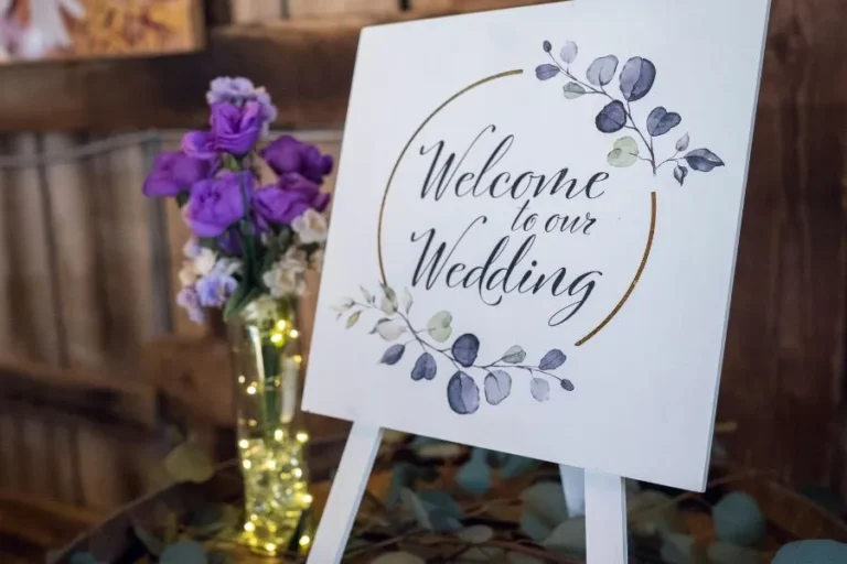 Cartel de bienvenida para boda decorado con hojas y flores moradas, sobre un caballete blanco elegante.