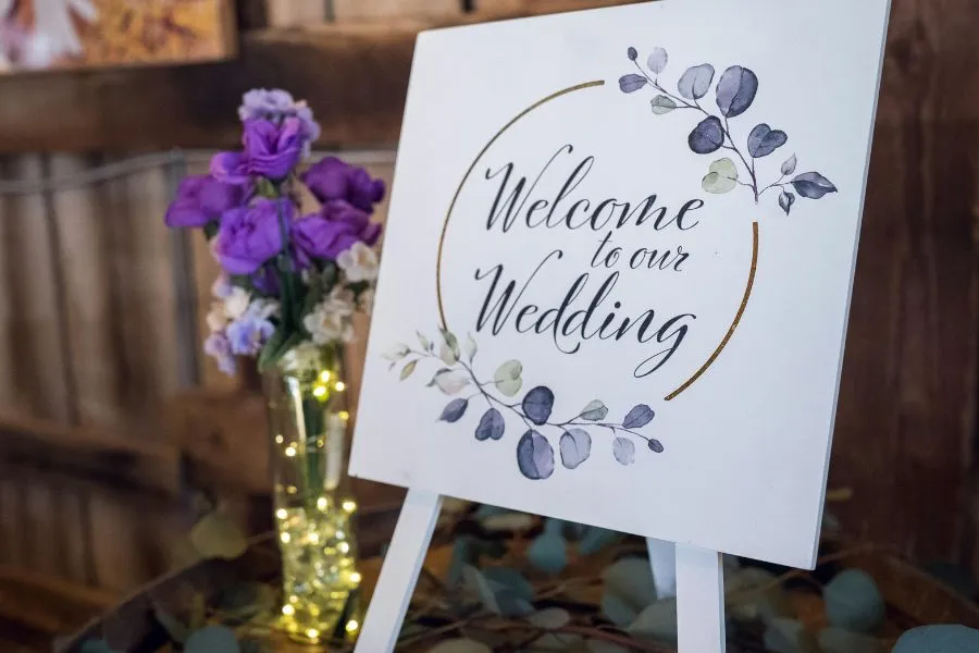 Cartel de bienvenida para boda decorado con hojas y flores moradas, sobre un caballete blanco elegante.