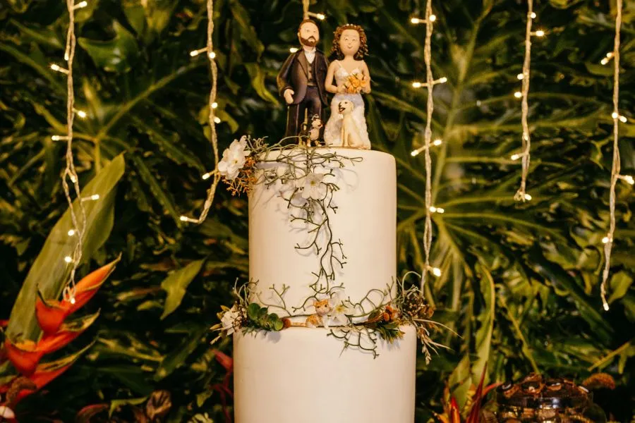 Figuras de novios encima de una tarta de dos pisos blanca con un fondo de selva